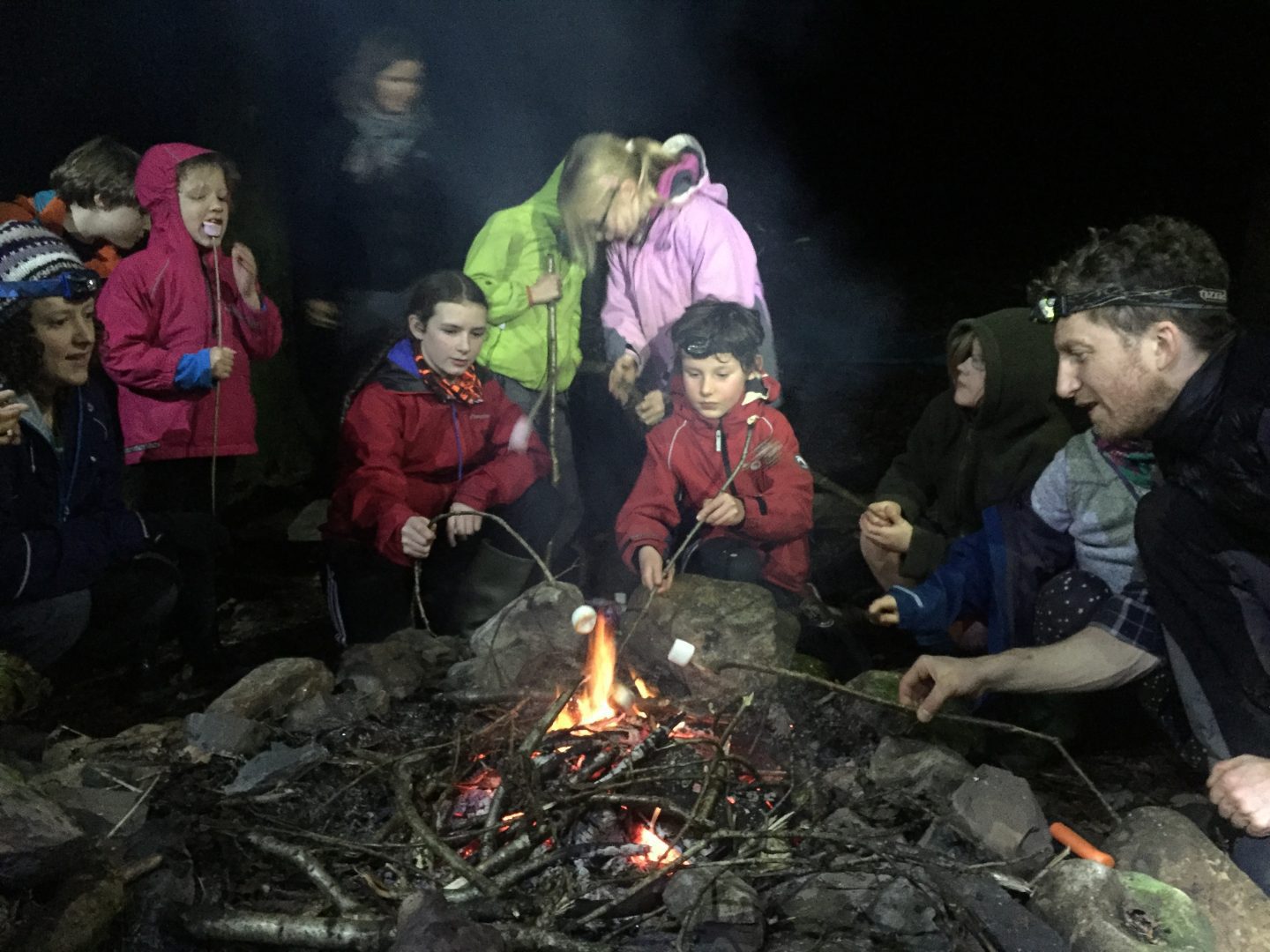 A group of adaults and children toasting marshmallows in the campfire