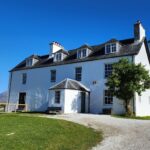Inchnadamph Lodge in assynt in the sunshine showing independent hostel sign and a lady on the decking