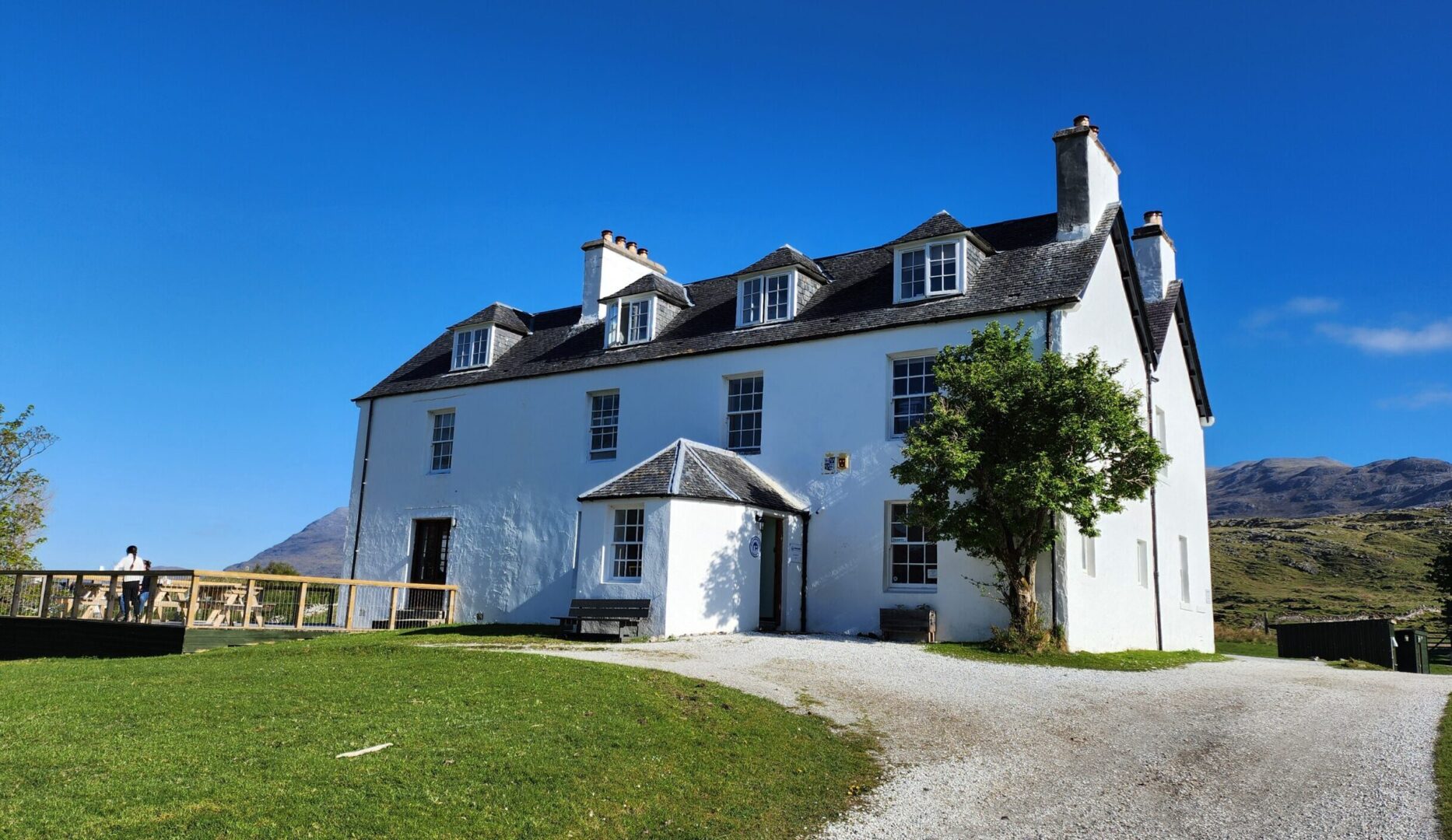 Inchnadamph Lodge in assynt in the sunshine showing independent hostel sign and a lady on the decking