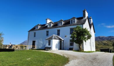 Inchnadamph Lodge in assynt in the sunshine showing independent hostel sign and a lady on the decking