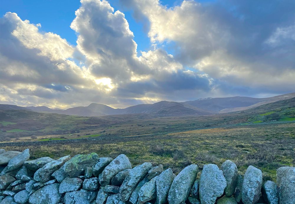 Landscape photo of the Snowdonian mountains taken from the Iron age hillfort. In the foreground is a cobblestone wall, and the shot spans out a grassy slope towards snowcapped peaks in the distance. The sky is a rich blue dappled with clouds and sunlight beaming through in stripes.