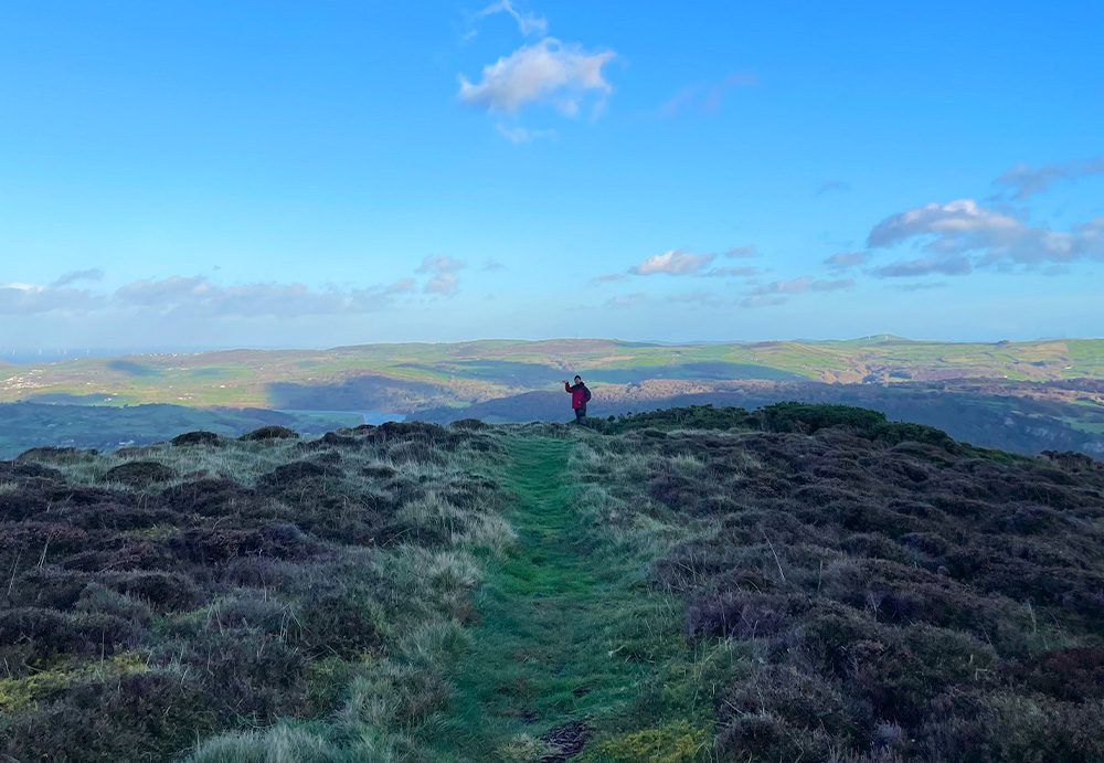 A distant figure in a red raincoat waves from the centre of the image, standing atop a grassy peak, blue sky in the distance and a few dappled clouds.