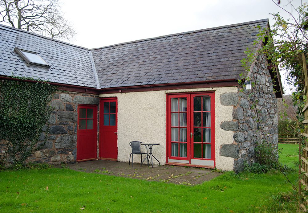 An exterior shot of the courtyard at Conwy Valley Barn. The double doors to this dorm room are painted red, with glass windows and curtains. There are 2 further doors into the common room and the second dorm. There is a small glass table and chairs in the courtyard.
