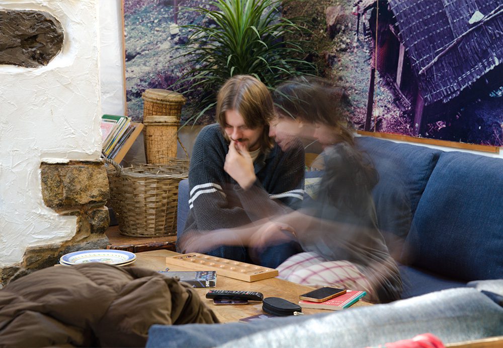 A long exposure photo of 2 people playing Mancala on a blue sofa, with books and board games in a pile on the shelf behind.