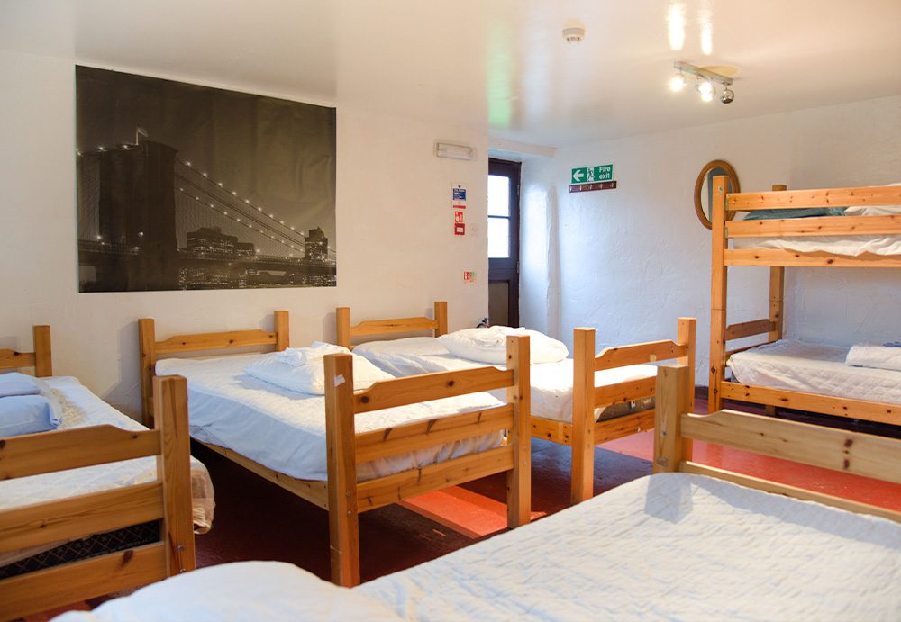 A photograph of one of the three dorm rooms at the site, 4 single beds and 1 bunkbed in view. The beds are oak, the carpet is red and the bed linen is white.