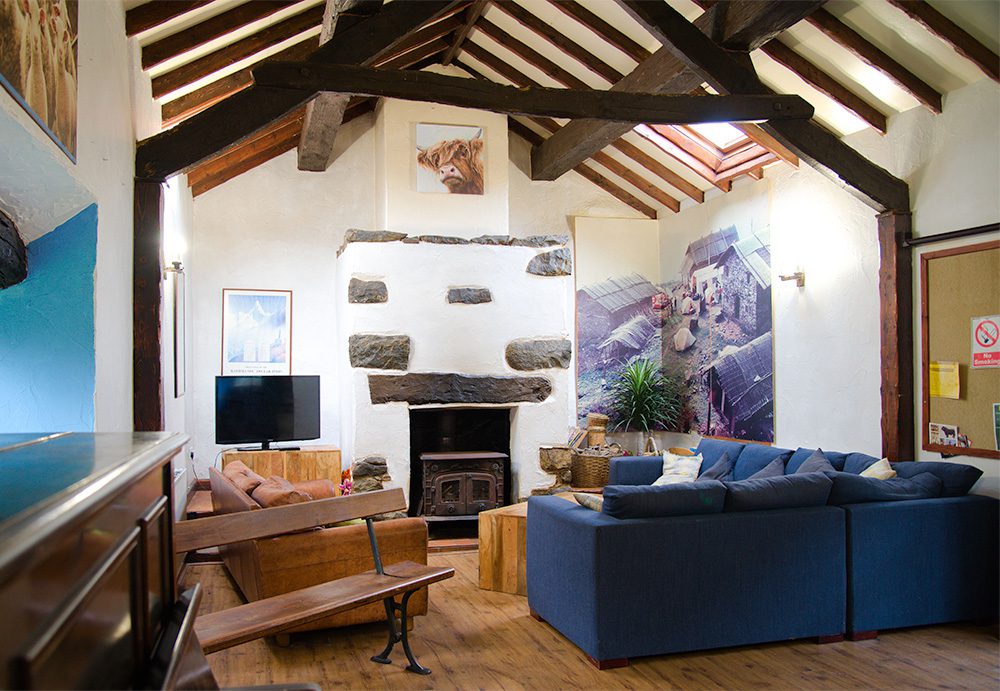 Wide angle photograph of the common room in Conwy Valley Barn, showing large blue L shaped sofa, brown leather sofa, TV and traditional hearth with a log burner. Ahead is a wooden beamed barn roof, and on the walls are photos of mountainous views and notice board with flyers.