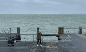 Person wearing a green raincoat standing on the end of a pier, with the stormy sea in the background