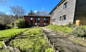 Two buildings, one brick and one wood, are placed at right angles with a fenced garden path joining them together. 