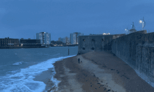 A beach in a city with stone military fort at the end, there is person walking a dog on the beach